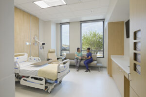 Nurse helps a patient in a hospital room. They look at a tablet together, seating on a small sofa in front of large windows.