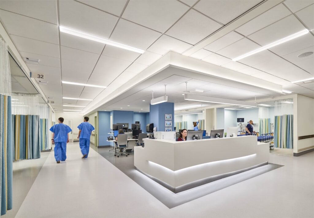 Nurses walking in a working room, with a desk and secretary. Curtains line the sectioned off hospital beds.